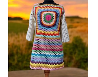 Puede incluir: Un vestido de crochet colorido con un patrón de rayas arcoíris y un gran diseño cuadrado de granny square en el centro. El vestido tiene mangas largas y un corte holgado.
