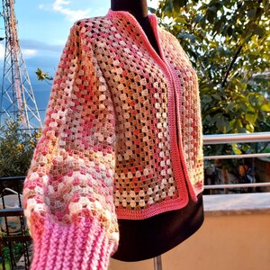 May include: A pink and brown crocheted cardigan with a button-up front. The cardigan has a granny square pattern and is made with a soft yarn.