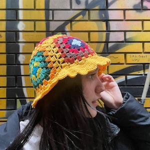 Op de afbeelding: Een kleurrijke gehaakte bucket hat met een gele rand en een veelkleurige bovenkant. De hoed is gemaakt met een granny square patroon en heeft verschillende kleuren, waaronder rood, oranje, geel, groen, blauw en paars.
