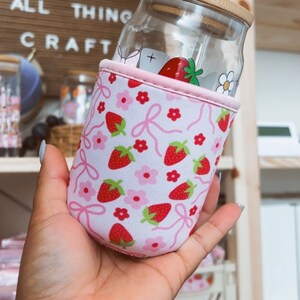 K&ouml;nnte beinhalten: Ein Glasgef&auml;&szlig; mit einem Holzdeckel und einer pink-wei&szlig;en Neoprenh&uuml;lle mit Erdbeermuster. Das Gef&auml;&szlig; hat einen durchsichtigen Glasstrohhalm, der aus dem Deckel herausragt. Das Gef&auml;&szlig; wird von einer Hand gehalten.
