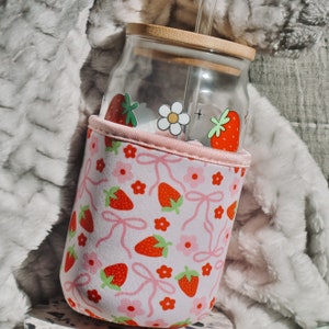 K&ouml;nnte beinhalten: Eine wiederverwendbare Glasflasche in Rosa und Wei&szlig; mit einem Strohhalm und einer pink-wei&szlig;en H&uuml;lle mit Erdbeer- und Blumenmuster.