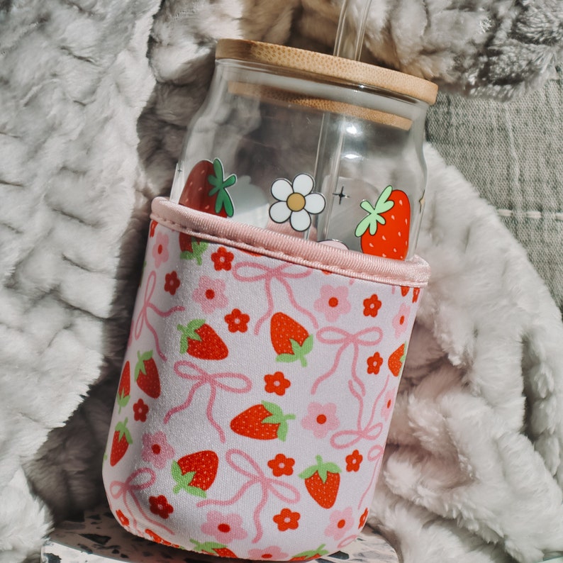 K&ouml;nnte beinhalten: Eine wiederverwendbare Glasflasche in Rosa und Wei&szlig; mit einem Strohhalm und einer pink-wei&szlig;en H&uuml;lle mit Erdbeer- und Blumenmuster.