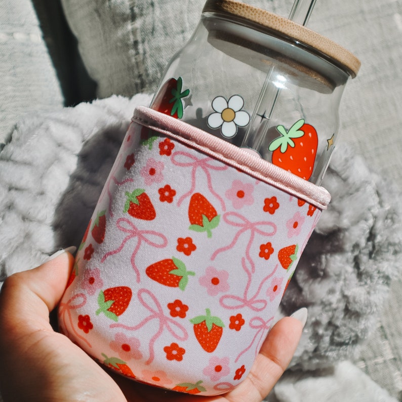K&ouml;nnte beinhalten: Eine rosa und wei&szlig;e isolierte Becherh&uuml;lle mit einem Erdbeer- und Blumenmuster. Die H&uuml;lle ist f&uuml;r eine Glasflasche mit Strohhalm konzipiert.