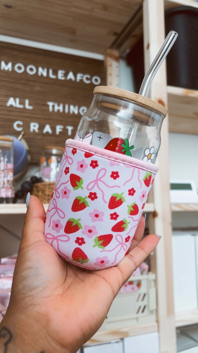 K&ouml;nnte beinhalten: Ein Glasgef&auml;&szlig; mit einem Holzdeckel und einer pink-wei&szlig;en Neoprenh&uuml;lle mit Erdbeermuster. Das Gef&auml;&szlig; hat einen durchsichtigen Glasstrohhalm, der aus dem Deckel herausragt. Das Gef&auml;&szlig; wird von einer Hand gehalten.