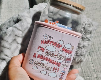Könnte beinhalten: Rosa isolierte Tasse mit einem weißen und rosa Design. Die Tasse hat den Text "Happiest when I'm reading & drinking iced coffee!!!" aufgedruckt.