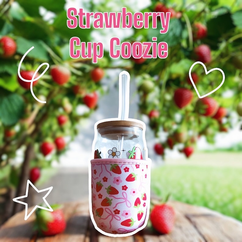 K&ouml;nnte beinhalten: Ein rosa-wei&szlig;er isolierter Becher-Coozie mit Erdbeer-Druck. Der Coozie befindet sich auf einem Glasgef&auml;&szlig; mit einem Strohhalm und einem Holzdeckel. Der Hintergrund ist ein verschwommenes Bild von Erdbeerpflanzen.