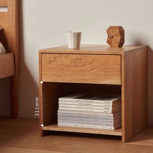 以下が含まれることがあります： 引き出しと棚が付いた木製ナイトスタンド。ナイトスタンドの高さは211mmで、ライトブラウンの仕上げ。棚には本が積み重ねられています。