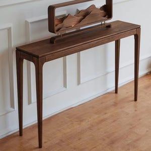 Op de afbeelding: Een donkerbruine houten console met een rechthoekig blad en vier slanke poten. Een decoratief houten stuk met een bergketenontwerp staat op het tafeloppervlak. De tafel staat tegen een witte muur.