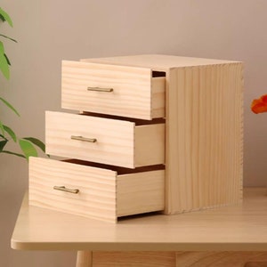 May include: A three-drawer wooden chest with a light wood finish and brass drawer pulls. The drawers are open, revealing the empty interior.