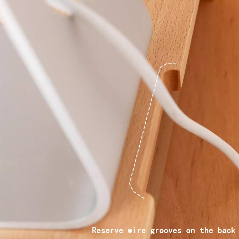 May include: A close-up of a wooden phone stand with a white phone charger cord running through a groove on the back. The stand has a slot for a phone and a groove for a cord.