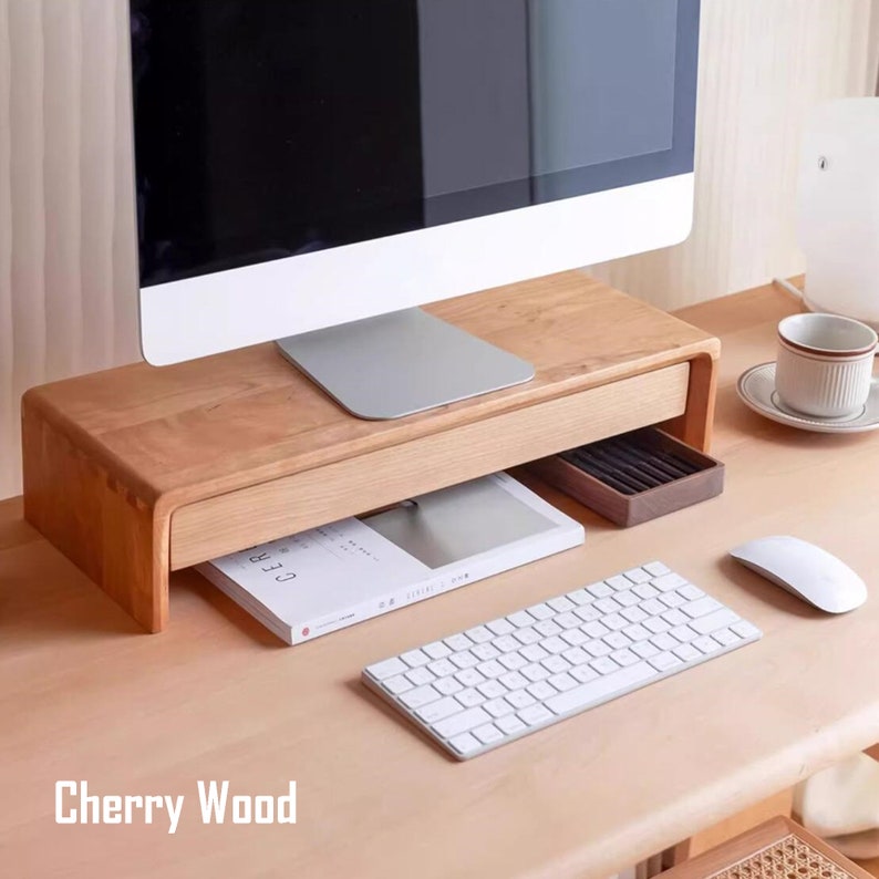 May include: A wooden monitor stand with a drawer for storage. The stand is made of cherry wood and has a natural finish. A white keyboard, a white mouse, and a book are on the desk in front of the stand. The text "Cherry Wood" is visible in the image.