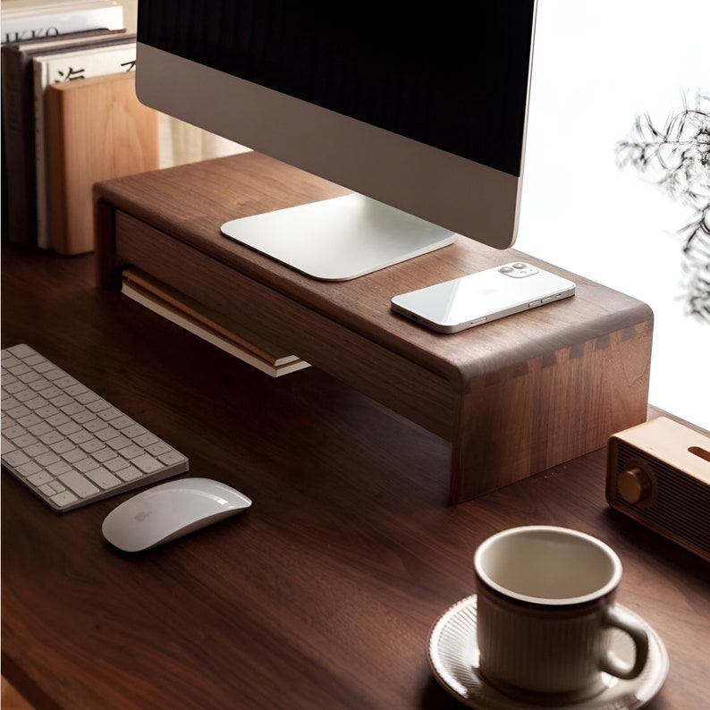 May include: A wooden monitor stand with a drawer for storage. The stand is made of walnut wood and has a natural finish. A computer monitor, a smartphone, and a keyboard are on the desk.