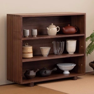 May include: A dark brown wooden bookshelf with three shelves, displaying various teacups, teapots, and serving bowls. The items are in neutral tones, including beige, brown, and white. The bookshelf is made of dark wood and has a simple, modern design.