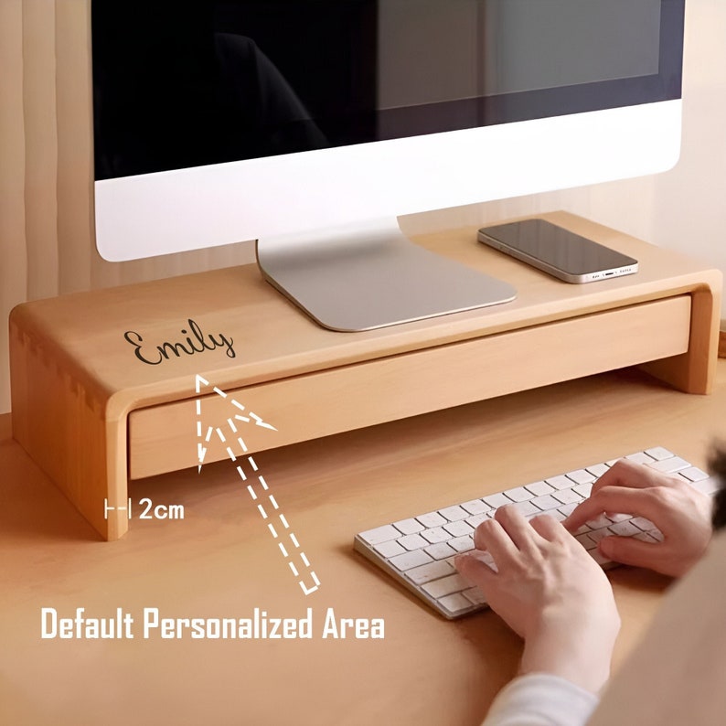 May include: A wooden monitor stand with a drawer. The stand is light brown and has a personalized area with the name "Emily" written on it. The drawer is 2 cm high.
