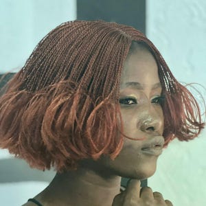 May include: A woman with reddish-brown micro braids styled in a bob. The braids are tightly woven, creating a textured appearance. The hair frames the face, with a slight gradient in color from darker roots to lighter ends. The person is wearing a black top.
