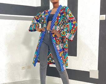 Op de afbeelding: Een kimono-achtige jas met lange mouwen. De jas heeft een levendige mix van rode, blauwe, groene en gele geometrische vormen. De jas wordt gedragen met een grijze jeans en zwarte hakken.