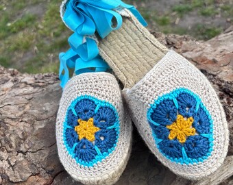 May include: A pair of espadrille sandals with a beige crochet upper and a woven jute sole. Each sandal features a blue and yellow floral design on the toe and is tied with bright blue ribbons.