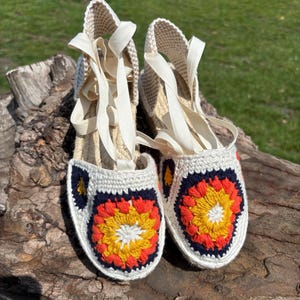 May include: A pair of handmade espadrille sandals with a crochet design. The sandals feature a white crochet upper with a navy blue, orange, and yellow floral pattern on the toe. They have beige jute soles and cream-colored ribbon ties.