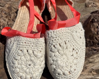 May include: A pair of off-white crochet espadrille sandals with coral-colored ribbon ties. The sandals have a woven jute sole and a floral crochet pattern on the toe. The ribbon ties wrap around the ankle.