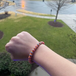 May include: A colorful beaded bracelet with orange, pink, blue, and gold beads. The bracelet is on a person's wrist.