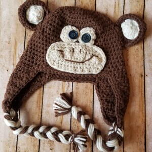 May include: Brown crocheted monkey hat with white yarn accents and black buttons for eyes. The hat has long brown and white braided ties.