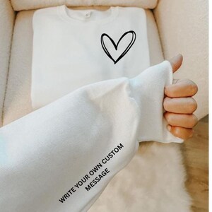 May include: White sweatshirt with a black heart graphic on the front. The sleeve displays the text "WRITE YOUR OWN CUSTOM MESSAGE". The sweatshirt is made of a soft material and is perfect for casual wear.