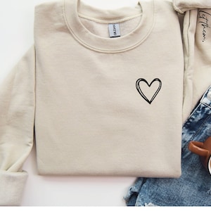 May include: A beige sweatshirt with a black hand-drawn heart on the front. The sweatshirt has a crew neck and long sleeves. The sleeves are rolled up. The sweatshirt is laying on a white surface with a pair of blue denim shorts and a brown mug with coffee.