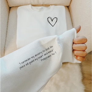 May include: White sweatshirt with a black heart on the front and a quote on the sleeve that reads "I surrender my anxiety to God. His peace will guard my heart and mind." Philippians 4:6-7