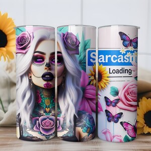 May include: Three stainless steel tumblers with different designs. The first tumbler features a woman with a sugar skull makeup design and floral tattoos. The second tumbler features a similar design with a woman with a sugar skull makeup design and floral tattoos. The third tumbler features a pink and white background with butterflies and a blue sign that says "Sarcasm Loading".