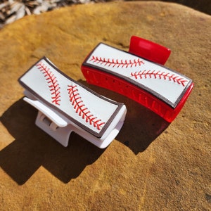 Two clips, one is white and the other red. Both are 4inch and have a leather piece on them that resemble a baseball. There is a brown border with white background and red stitches
