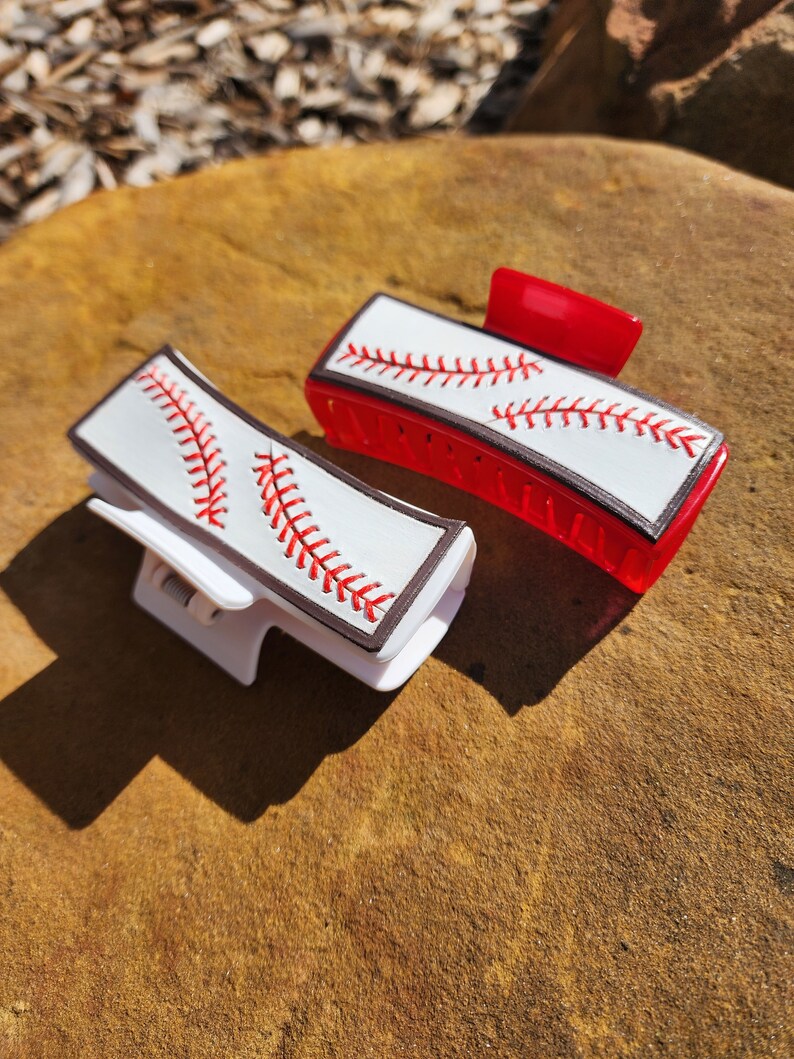 Two clips, one is white and the other red. Both are 4inch and have a leather piece on them that resemble a baseball. There is a brown border with white background and red stitches