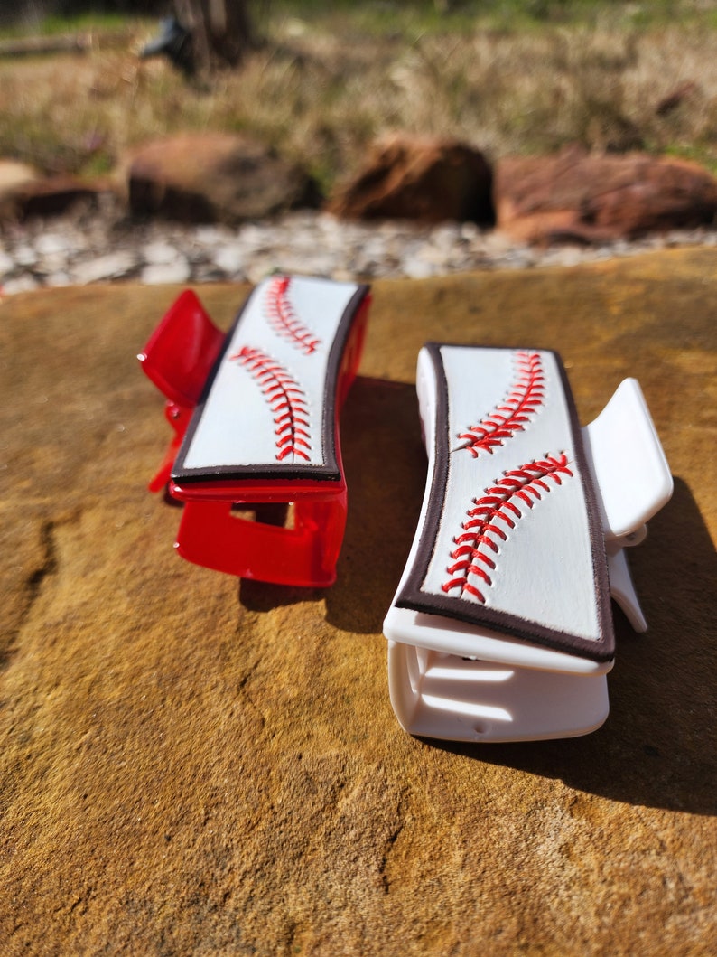 Two clips, one is white and the other red. Both are 4inch and have a leather piece on them that resemble a baseball. There is a brown border with white background and red stitches