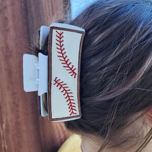 A 4inch white claw clip with leather piece on top of it that resembles a baseball. There is a brown border with white background and red stitches. The clip is shown being used in a full updo.