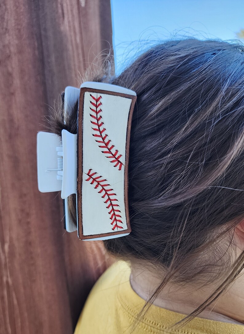 A 4inch white claw clip with leather piece on top of it that resembles a baseball. There is a brown border with white background and red stitches. The clip is shown being used in a full updo.