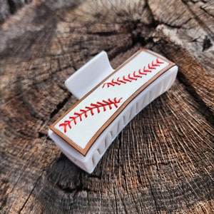 A 4inch white claw clip with leather piece on top of it that resembles a baseball. There is a brown border with white background and red stitches