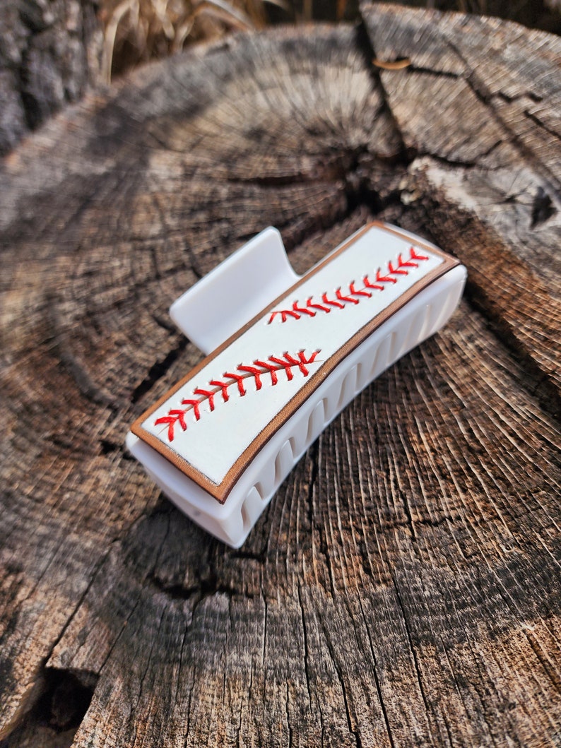 A 4inch white claw clip with leather piece on top of it that resembles a baseball. There is a brown border with white background and red stitches