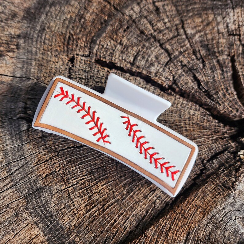 A 4inch white claw clip with leather piece on top of it that resembles a baseball. There is a brown border with white background and red stitches