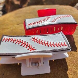Two clips, one is white and the other red. Both are 4inch and have a leather piece on them that resemble a baseball. There is a brown border with white background and red stitches