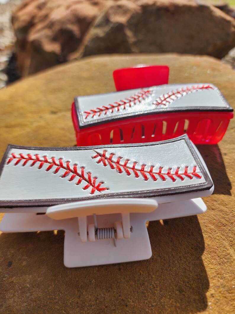 Two clips, one is white and the other red. Both are 4inch and have a leather piece on them that resemble a baseball. There is a brown border with white background and red stitches