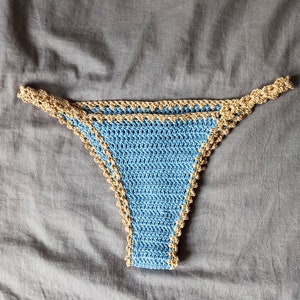 Peut inclure: Bas de bikini bleu clair en crochet, bordé de beige. Le travail au crochet est visible. Le vêtement est présenté sur un fond de tissu gris. Le bas de bikini a un design classique et minimaliste.