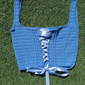 Peut inclure: Un haut crocheté bleu avec un devant à lacets et un ruban bleu assorti noué en un nœud. Le haut est posé sur une surface herbeuse verte.