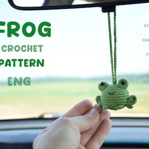 May include: A green crocheted frog car charm or plushie. The frog has black eyes and a smiling mouth. The text "FROG CROCHET PATTERN ENG" is in the background. The charm is hanging from a green string.