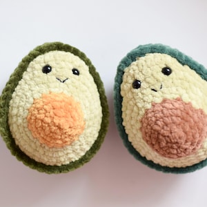 May include: Two plush avocado toys, each with a smiling face. One has a green exterior and an orange pit, the other has a teal exterior and a brown pit. The toys are made of a soft, textured material.