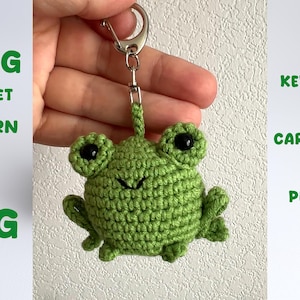 May include: A green crochet frog keychain with black eyes and a smiling mouth. The frog is attached to a silver-colored metal clasp. Text on the left reads "FROG CROCHET PATTERN ENG". Text on the right reads "KEYCHAIN CAR CHARM PLUSHIE".
