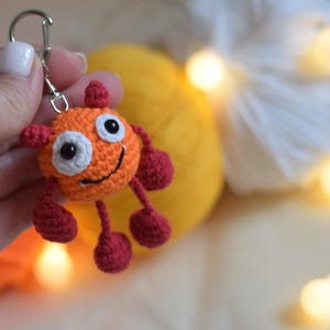 Peut inclure: Un porte-clés crocheté à la main en forme de monstre orange souriant avec des détails rouges. Il est doté d'un fermoir argenté. En arrière-plan, des pelotes de laine jaunes et blanches et une guirlande lumineuse.