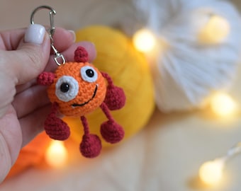 May include: A handmade orange crochet keychain in the shape of a smiling monster with red accents. The keychain has a silver clasp. The background includes yellow and white yarn balls and a string of lights.
