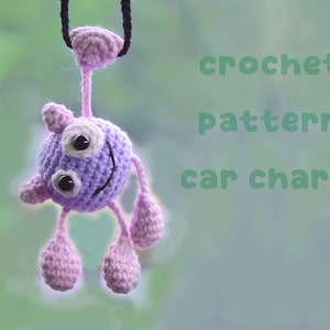 May include: A handmade crochet car charm in the shape of a friendly monster. The charm is primarily purple with pink accents, including the arms, legs, and details. The text "crochet pattern car charm" is visible in the image.