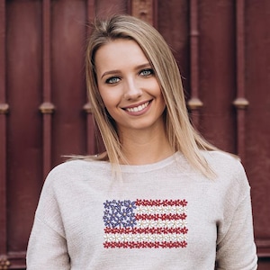 以下が含まれることがあります： アメリカ国旗のデザインが刺繍された、明るい灰色のスウェットシャツを着た女性。旗は赤、白、青の花でできています。背景は濃い赤色のドアです。