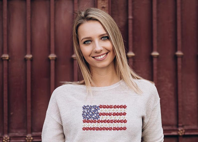以下が含まれることがあります： アメリカ国旗のデザインが刺繍された、明るい灰色のスウェットシャツを着た女性。旗は赤、白、青の花でできています。背景は濃い赤色のドアです。