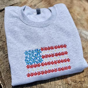 以下が含まれることがあります： 刺繍されたアメリカ国旗のデザインが施された、明るい灰色のスウェットシャツ。旗の星は青い花で、ストライプは赤と白の花でできています。スウェットシャツは柔らかい素材で作られています。